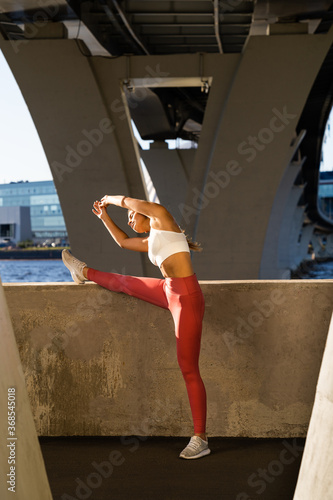 Slim sportswoman in pink legging standing under the bridge, stretching muscles making functional training, does warm-up outdoors, exercises for the back muscles. 
