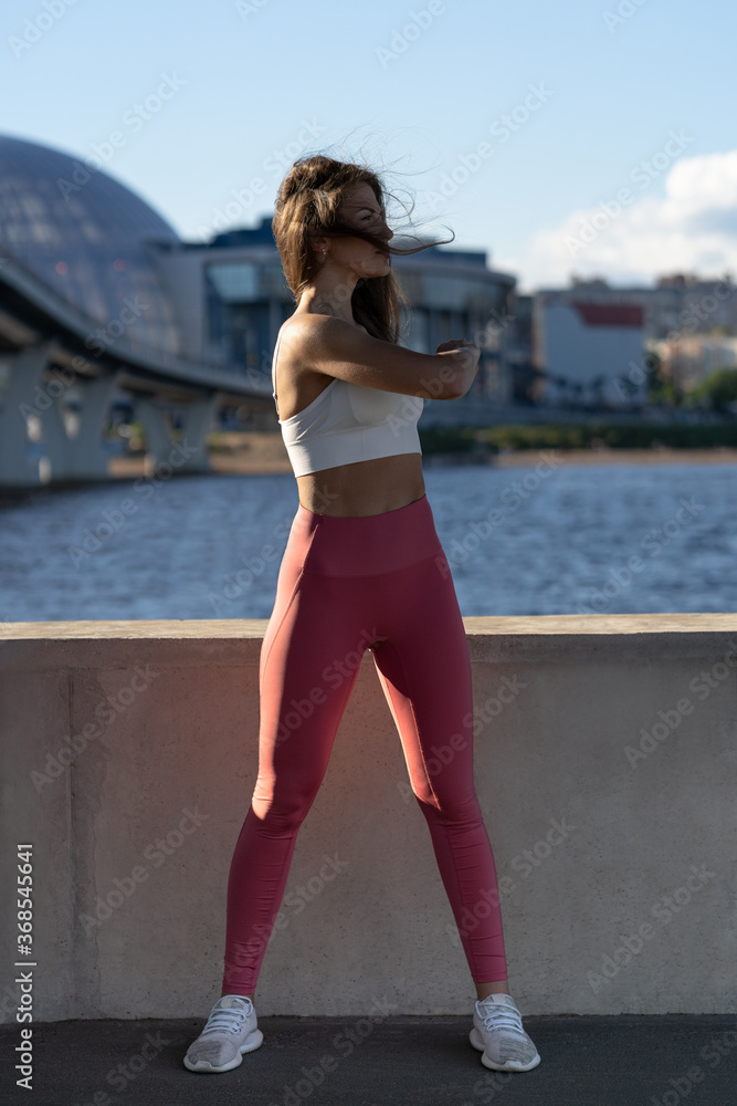 Fit young athletic woman stretching arms and shoulder joints on embankment. Fitness female in pink leggings doing warmup workout outdoor. 