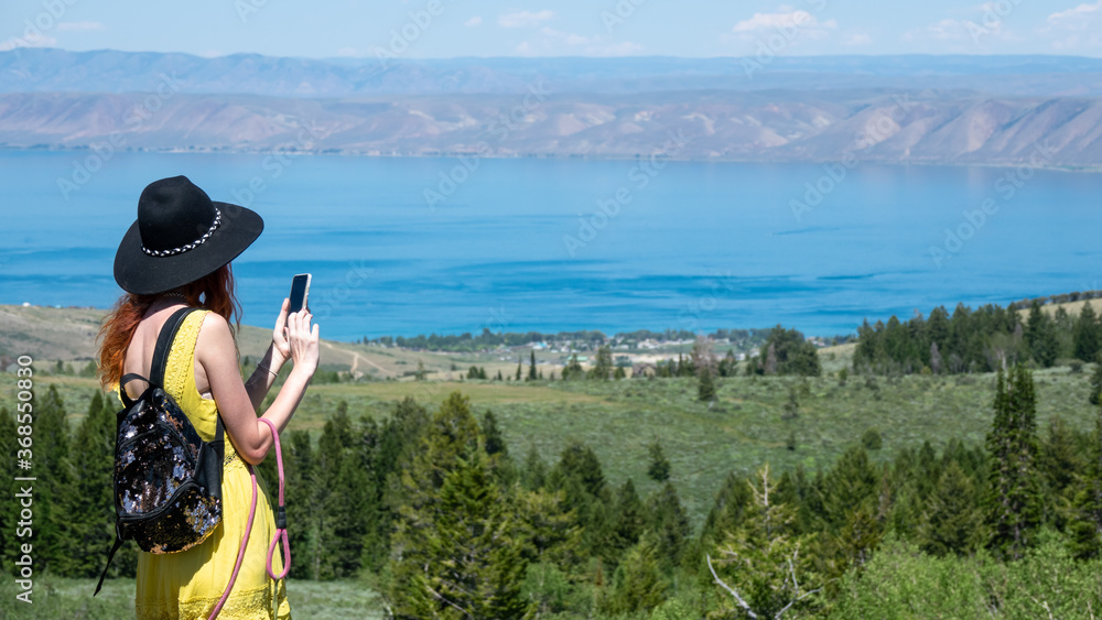Obraz premium Young Girl with yellow dress and black hat taking photo of Lake Utah View point