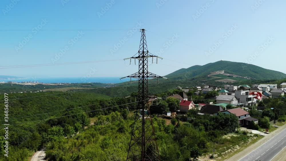 Aerial shot of an electric tower in the countryside among green mountains. The blue sky over the village is connected to electrical wires through a special device. Slow boom down.