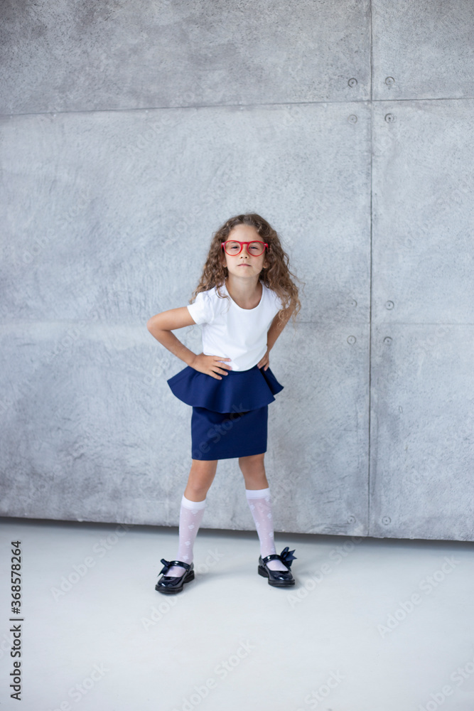 Vertical front photo, full size, with a gorgeous little girl, looking good, in a skirt, with her hands on her waist in red glasses, isolated on the background. Vertical front photo, full size, with a gorgeous little girl, looking good, in a skirt, with her hands on her waist in red glasses, isolated on the background.