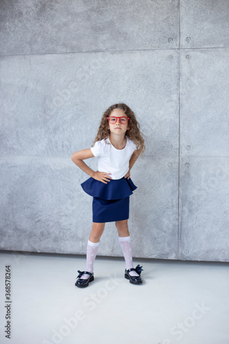 Vertical front photo, full size, with a gorgeous little girl, looking good, in a skirt, with her hands on her waist in red glasses, isolated on the background.