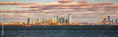 A sunset panorama of Jersey City skyline featuring the skyscrapers, upper bay, Hudson river mouth, part of Manhattan and the statue of liberty. Sunlight reflects from the surface of the buildings. © Grandbrothers