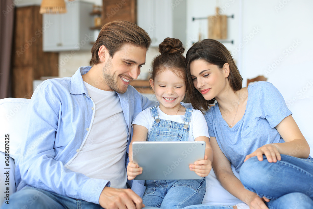 Positive friendly young parents with smiling little daughter sitting on sofa together answering video call on digital tablet while relaxing at home on weekend. Positive friendly young parents with smiling little daughter sitting on sofa together answering video call on digital tablet while relaxing at home on weekend.