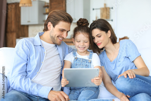 Positive friendly young parents with smiling little daughter sitting on sofa together answering video call on digital tablet while relaxing at home on weekend.