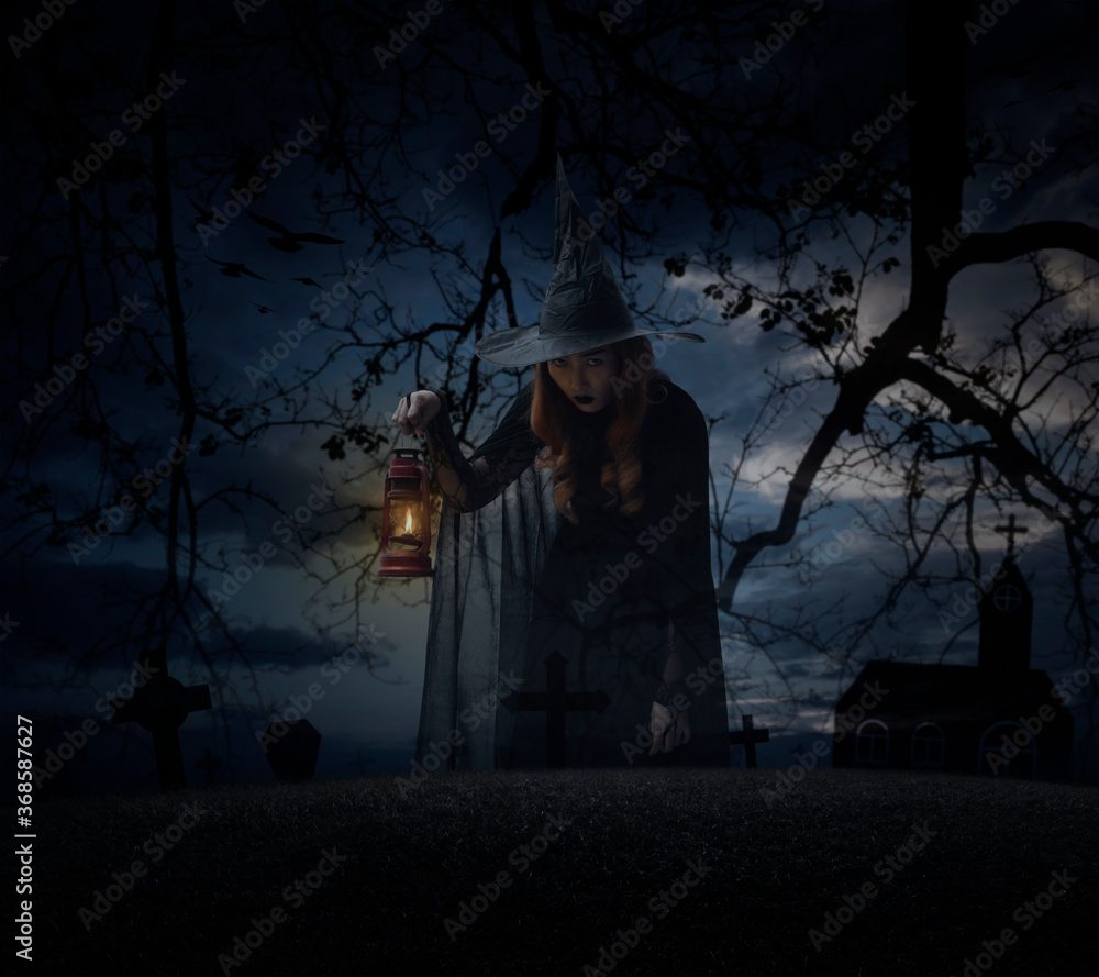 Halloween witch holding ancient lamp standing over grass, dead tree, cross, birds with church over spooky cloudy sky, Halloween mystery concept
