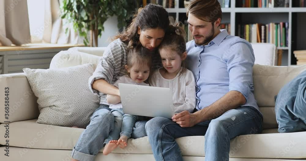 Happy young couple parents showing educational application on computer to little cute children daughters at home, early education concept. Smiling mum and dad watching cartoons with kids on laptop. Happy young couple parents showing educational application on computer to little cute children daughters at home, early education concept. Smiling mum and dad watching cartoons with kids on laptop.