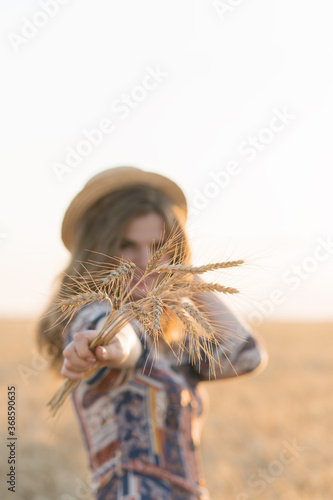 Beautiful young woman holding a bunch of wheat in her hands, in a wheat field on a sunset background. Selective focus