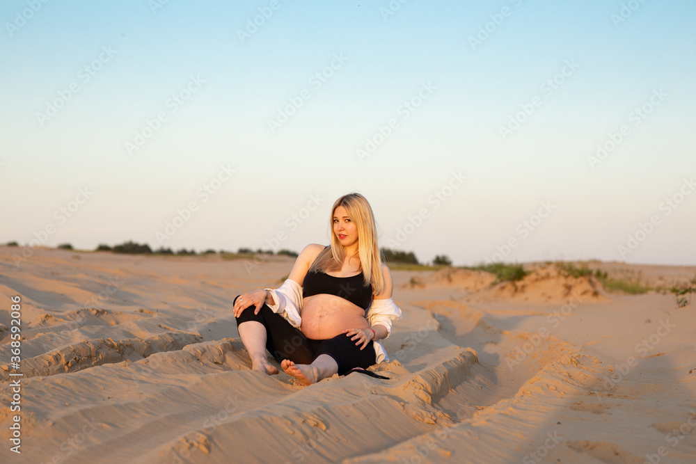 Young pregnant woman with long blondy hair wearing a black top sitting in a desert among the sand in the evening Young pregnant woman with long blondy hair wearing a black top sitting in a desert among the sand in the evening