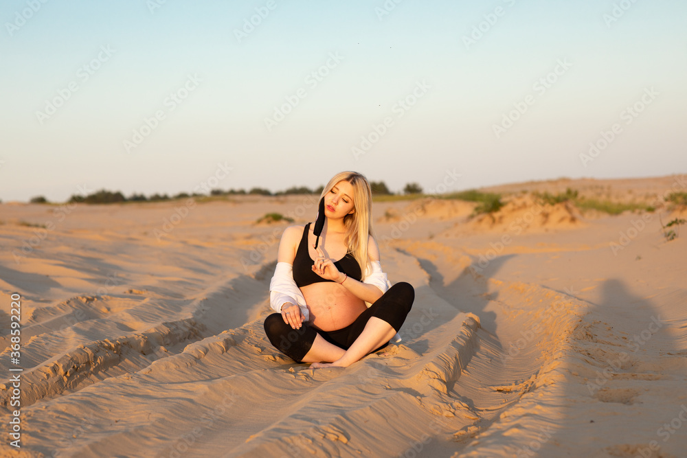 Young pregnant woman with long blondy hair wearing a black top sitting in a desert among the sand in the evening Young pregnant woman with long blondy hair wearing a black top sitting in a desert among the sand in the evening