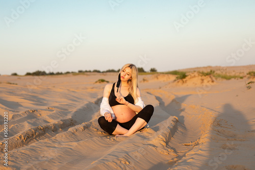 Young pregnant woman with long blondy hair wearing a black top sitting in a desert among the sand in the evening 