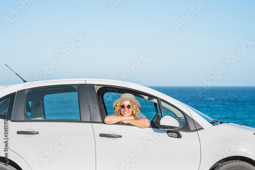 one beautiful happy woman looking at the camera from inside of a car with the sea or ocean at the background - adult traveler and nomad lifestyle and concept - outdoor sunny day