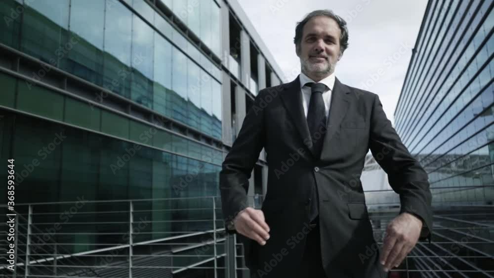 Successful mature businessman in office suit and tie looking at camera, smiling, making arms crossed while standing between city buildings. Front view, low angle. Male portrait concept Successful mature businessman in office suit and tie looking at camera, smiling, making arms crossed while standing between city buildings. Front view, low angle. Male portrait concept