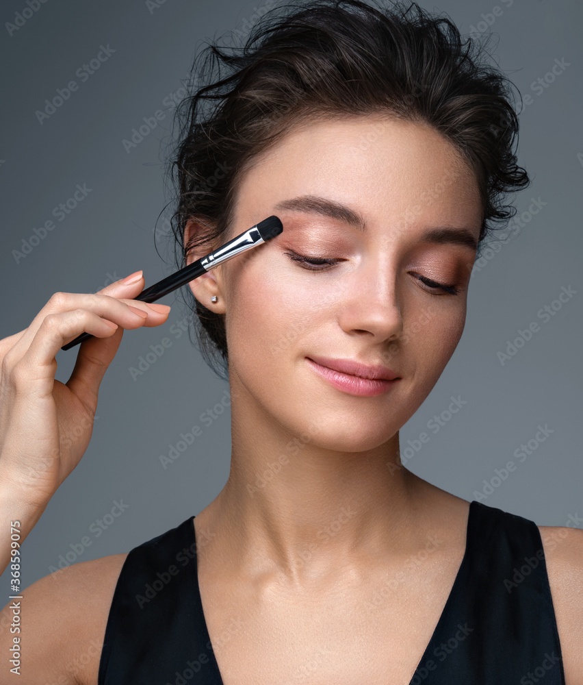 © Romario Ien - Beautiful woman using brush for eyeshadow. Photo of woman with perfect makeup on gray background. Beauty concept