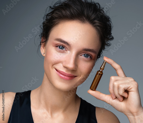 Woman holds ampoule with serum for hair or skin care. Photo of attractive woman with perfect makeup on gray background. Beauty concept photo