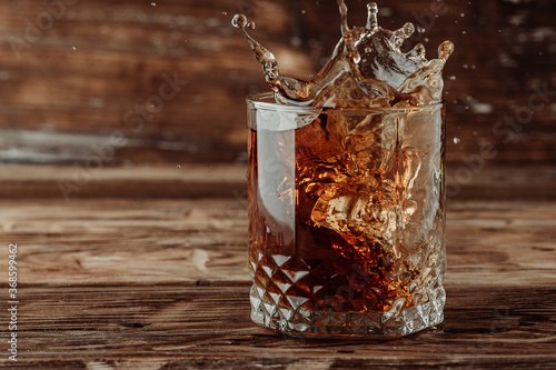 Frozen movement of a splash of Whiskey cognac or brandy in a beautiful glass due to a thrown ice cube. Wooden rustic background with a monochromatic brown tone. photo
