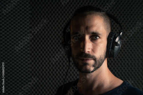 Hombre con peinado moderno y ojos azules, y camiseta gris de mangas azul marino con auriculares en un estudio de grabación de sonido mirando a cámara con luz de flash