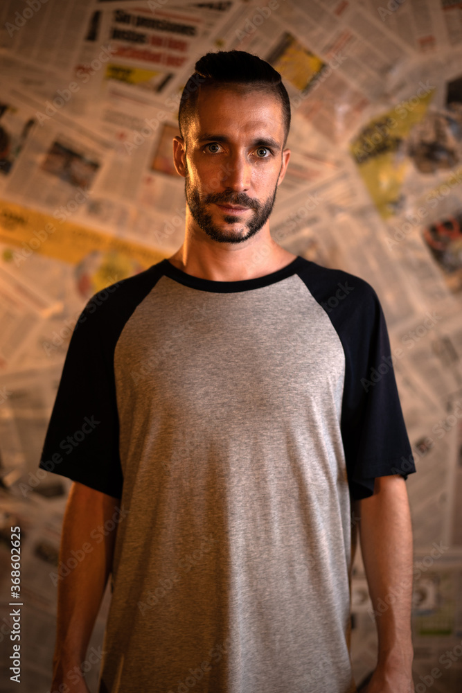 Hombre con peinado moderno y ojos azules, y camiseta gris de mangas azul marino en fondo de papel de periódico, mirando a cámara con rostro serio Hombre con peinado moderno y ojos azules, y camiseta gris de mangas azul marino en fondo de papel de periódico, mirando a cámara con rostro serio