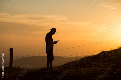 Portrait of man silhouette standing at the top of the mountain looking his smartpnone in hands on sunset background