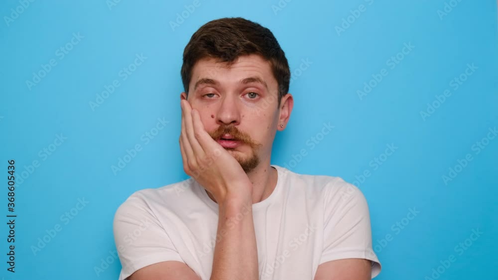 Exhausted young man feeling bored, lack of motivation, apathy, wears t-shirt, isolated on blue studio background. Sad guy with frowning expression, looks unhappy straight at camera. Human emotions Exhausted young man feeling bored, lack of motivation, apathy, wears t-shirt, isolated on blue studio background. Sad guy with frowning expression, looks unhappy straight at camera. Human emotions