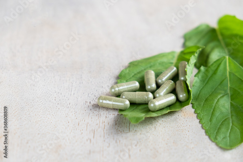Selective focus at Herbal medicine in capsule on brown wood surface, with green  holy basil leaf herb at the background. Healthcare and medical lifestyle concept.