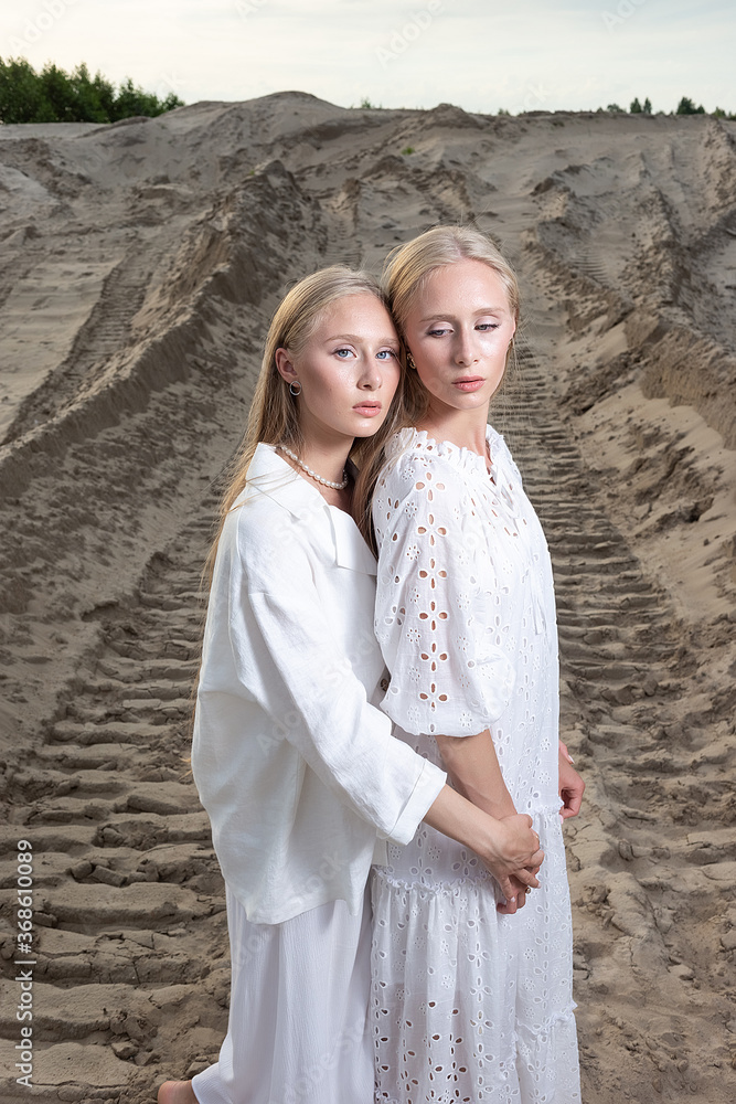 young pretty twins with long blond hair embrace at sand quarry in elegant white dress, skirt, jacket. stylish fashion photoshoot, summer photosession. identical sisters spend time together outdoors