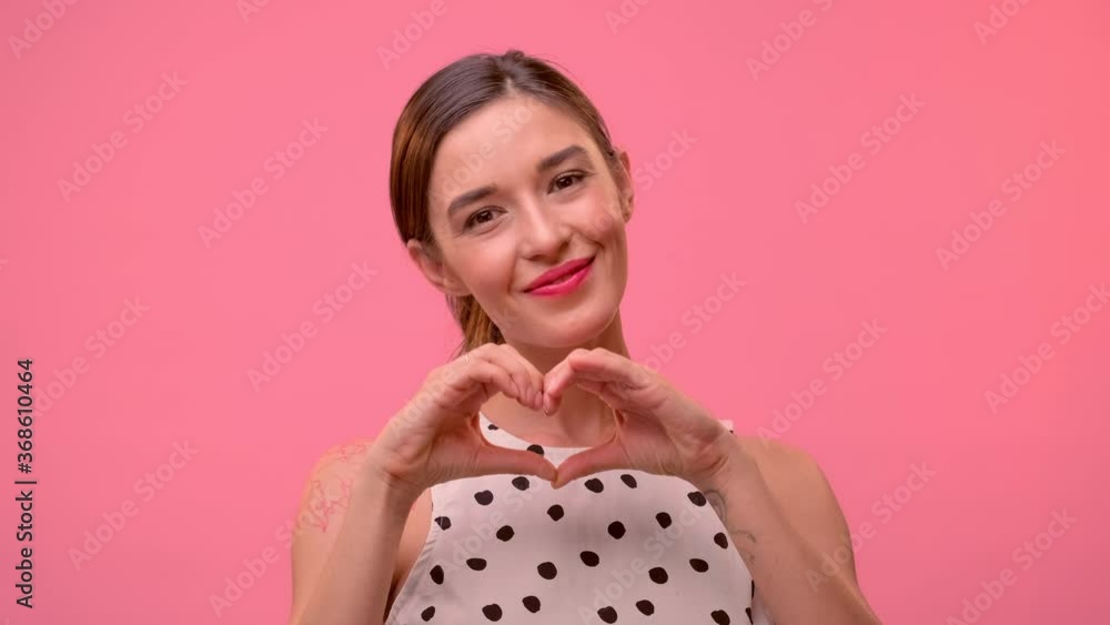 Attractive European female makes heart shape gesture, expresses love, says be my valentine, smiles positively, wears white dress, poses against pink background. Body language concept Attractive European female makes heart shape gesture, expresses love, says be my valentine, smiles positively, wears white dress, poses against pink background. Body language concept