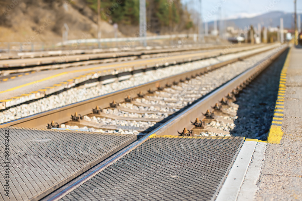 Empty train station in small city on sunny day, shallow depth of field photo, focus on concrete platform, passenger crossing, and steel rail foreground