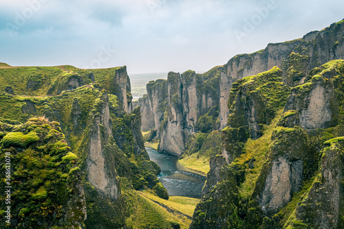 View of Fjaðrárgljúfur canyon in Iceland, with river and waterfall nearby, green grass and beautiful sky.