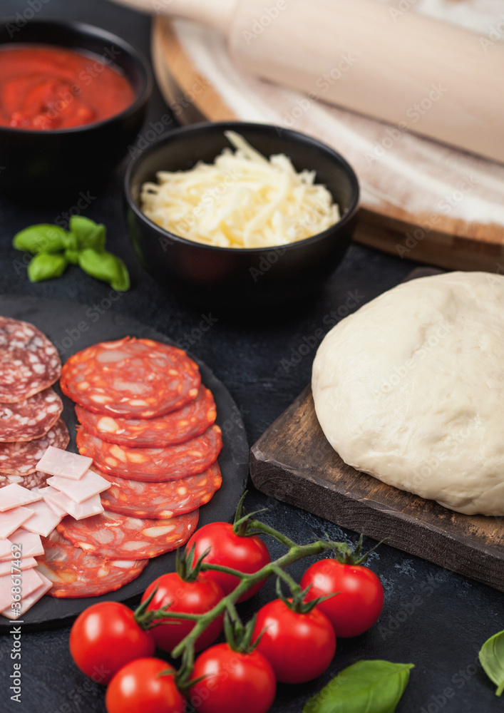 Preparation for baking of pepperoni pizza with raw dough, salami spicy chorizo with wheel cutter and fresh tomatoes and basil on dark background with bowl plates with cheese and tomato paste. Preparation for baking of pepperoni pizza with raw dough, salami spicy chorizo with wheel cutter and fresh tomatoes and basil on dark background with bowl plates with cheese and tomato paste.