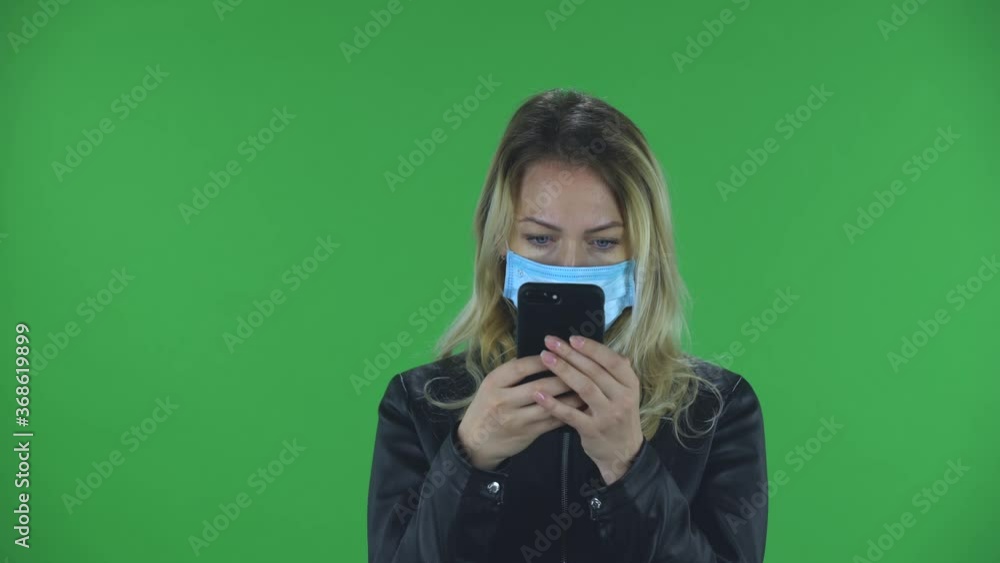 Portrait of beautiful young woman in medical mask is texting on her phone. Blonde with loose hair in a black jacket and jeans on a green screen in the studio. Health Protection Corona Virus Concept. Portrait of beautiful young woman in medical mask is texting on her phone. Blonde with loose hair in a black jacket and jeans on a green screen in the studio. Health Protection Corona Virus Concept.