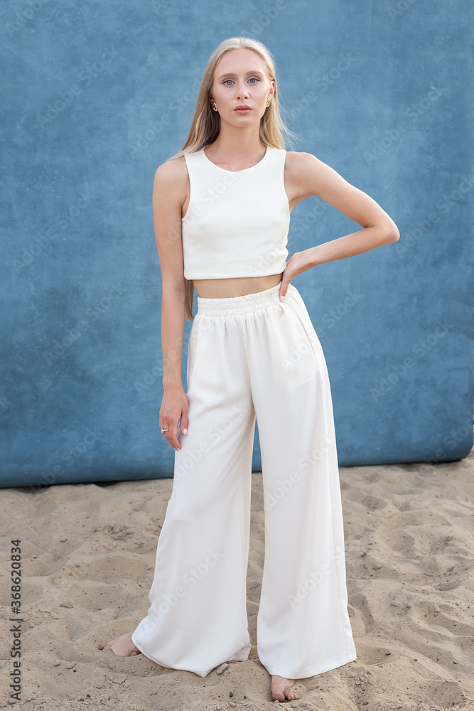 Fototapeta premium beauty portrait outdoors on sand in front of blue background, pretty female posing. attractive caucasian girl with long hair looking at camera. beautiful woman in white clothes, elegant fashion style