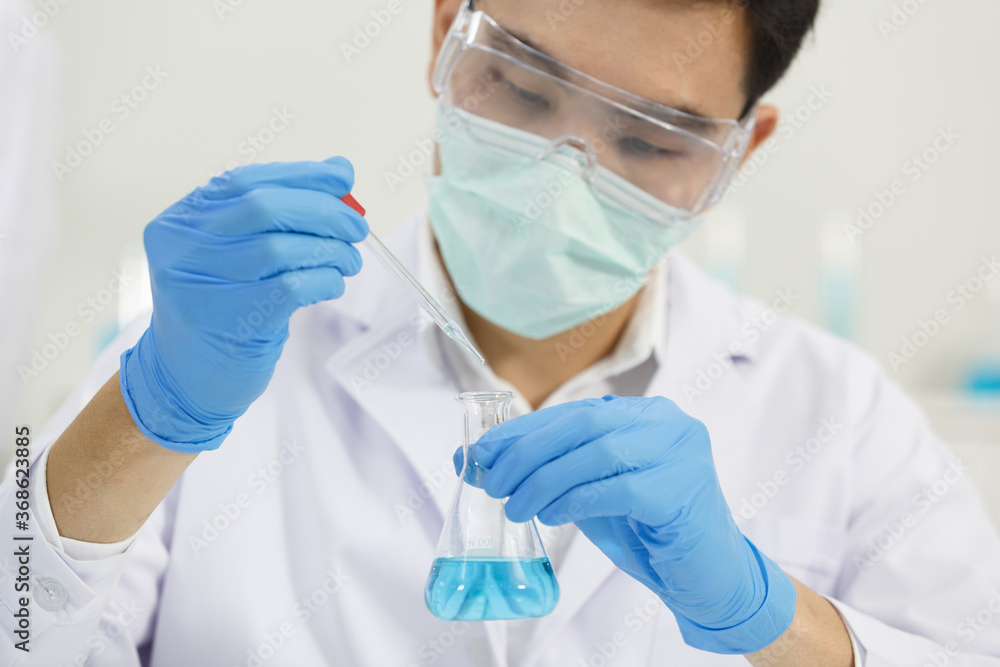 Male Asian Scientist wears Mask working in Lab while Droping Solution to Erlenmeyer Flask. Blue Tone. SARS-CoV-2 , Covid-19 THEME. Male Asian Scientist wears Mask working in Lab while Droping Solution to Erlenmeyer Flask. Blue Tone. SARS-CoV-2 , Covid-19 THEME.