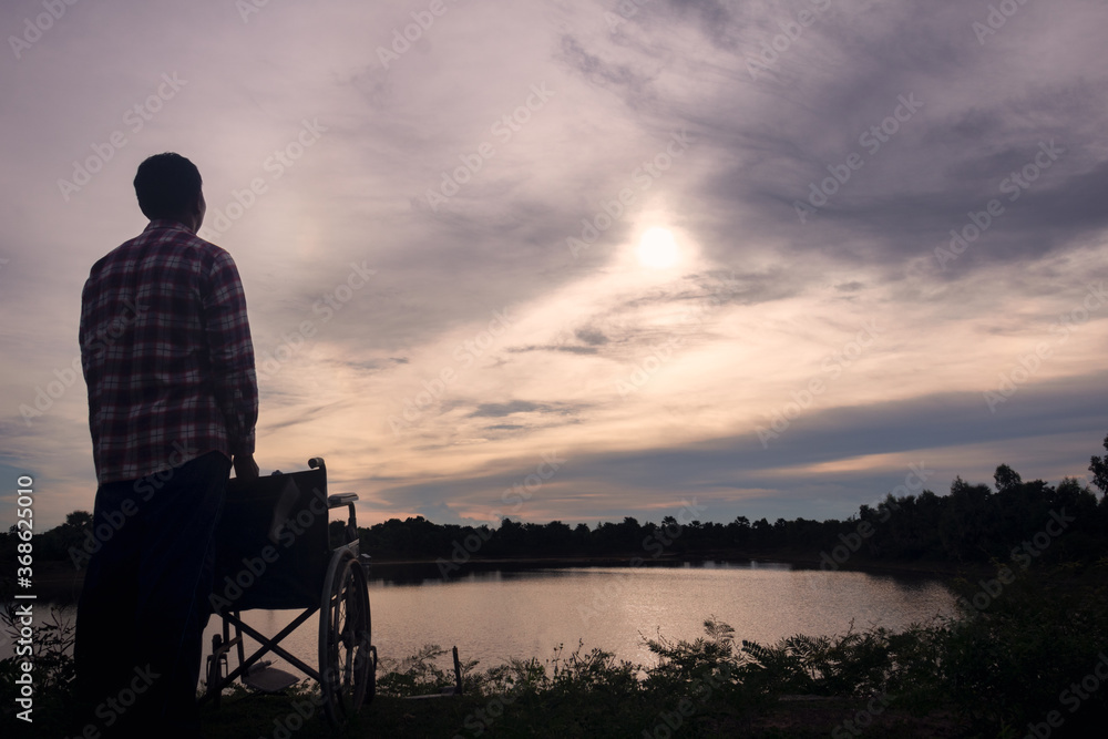 Fototapeta premium Silhouette young disabled man seeing river background.He is raise a hand to and sitting on wheelchair.despair,lonely,hope.Photo concept depression and Patient.