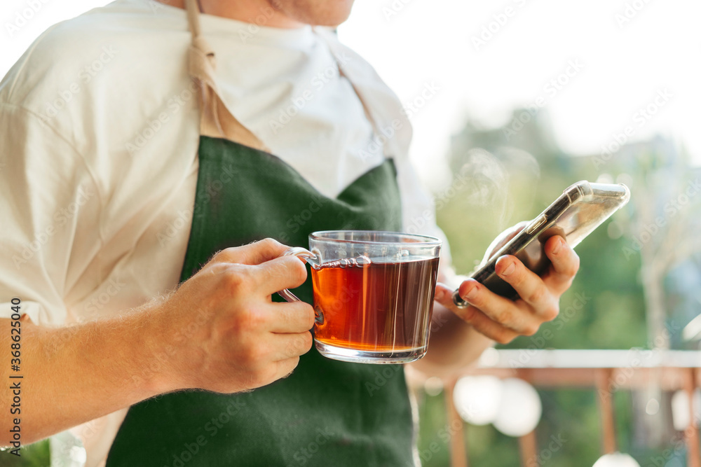 Crop close-up man drinks hot tea and holds a smartphone in his hands on a sunny day. Business success. Stay at home concept. Crop close-up man drinks hot tea and holds a smartphone in his hands on a sunny day. Business success. Stay at home concept.