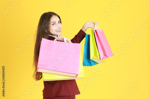 Beautiful working woman in red suit smile, happy and hand holding colorful shopping paper bags isolated on yellow background.