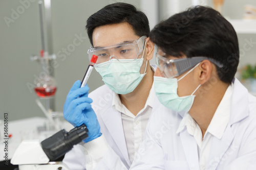 Two Male Scientists wear Face Mask working in Lab while Checking Blood tested by micro scope. SARS-CoV-2 , Covid-19 THEME.