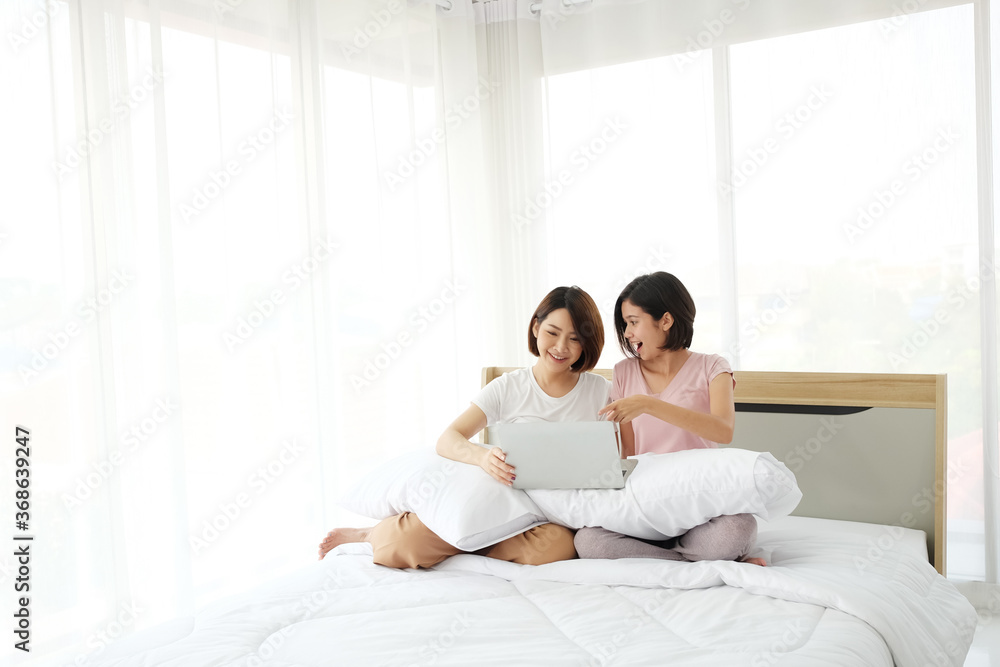 Asian young sisters lovely couple sitting on white bed and smiling together for online Social with laptop in bedroom. Technology and Homosexual women or Lesbian lifestyle concept.