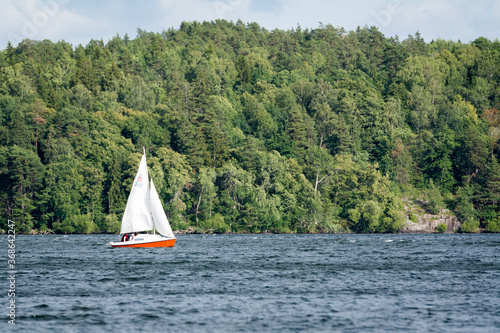 Small red sailboat sailing on a lake near Stockholm in front of green trees on a sunny day.