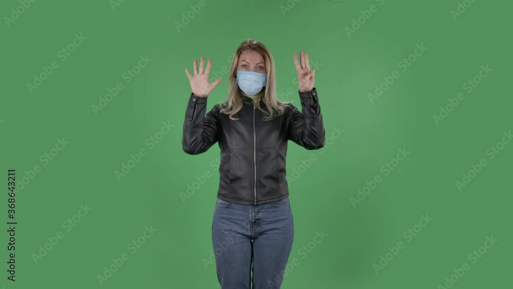 Portrait of beautiful young woman in medical protective face mask is looking at camera and shows fist, then counting one, two, three, four, five fingers. Blonde with loose hair in a black jacket and Portrait of beautiful young woman in medical protective face mask is looking at camera and shows fist, then counting one, two, three, four, five fingers. Blonde with loose hair in a black jacket and
