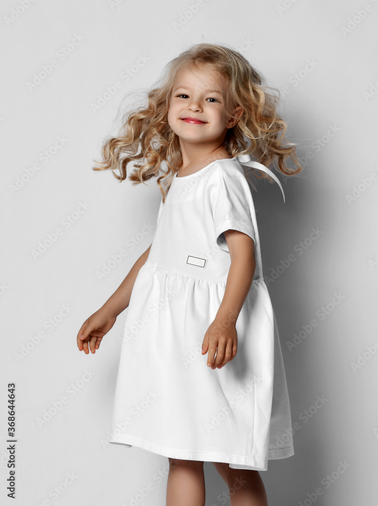 Little blonde curly positive princess girl in white casual dress and sneakers standing walking with curly hair over grey wall Little blonde curly positive princess girl in white casual dress and sneakers standing walking with curly hair over grey wall