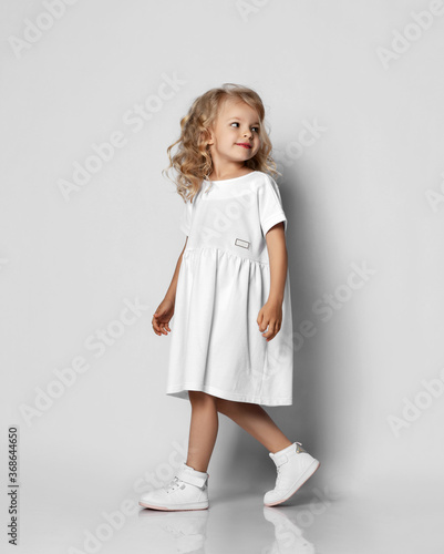 Little blonde curly positive princess girl in white casual dress and sneakers standing walking with curly hair over grey wall