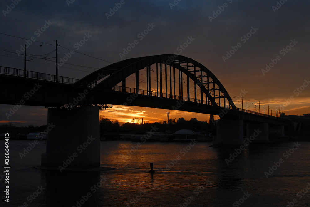 The Old Sava Bridge at sunset in Belgrade, Serbia. The bridge crosses the Sava River in central Belgrade. The bridge is silhouetted against the orange glow of the sunset.