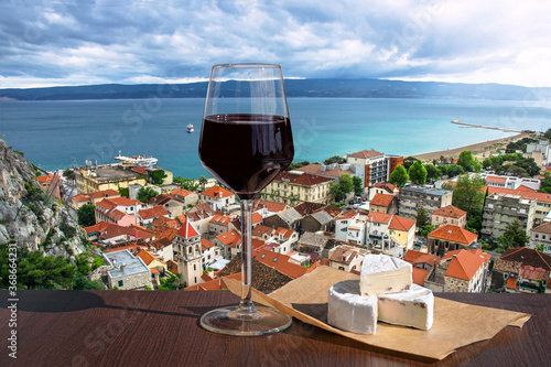 Fototapeta Naklejka Na Ścianę i Meble -  Glass of red wine with brie cheese against view of the small town Omis surrounded with mountains, Cetina river and sea, Makarska Riviera, Croatia. View of pine tree,  old city center with red roofs.