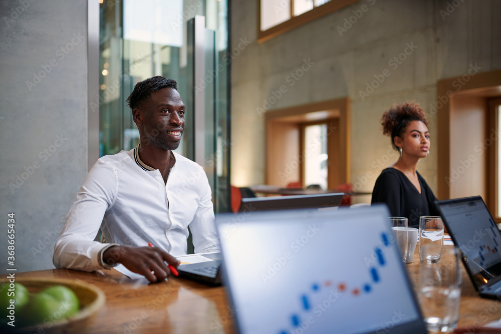 Smiling businessman looking away while sitting by female colleague at creative workplace during meeting Smiling businessman looking away while sitting by female colleague at creative workplace during meeting