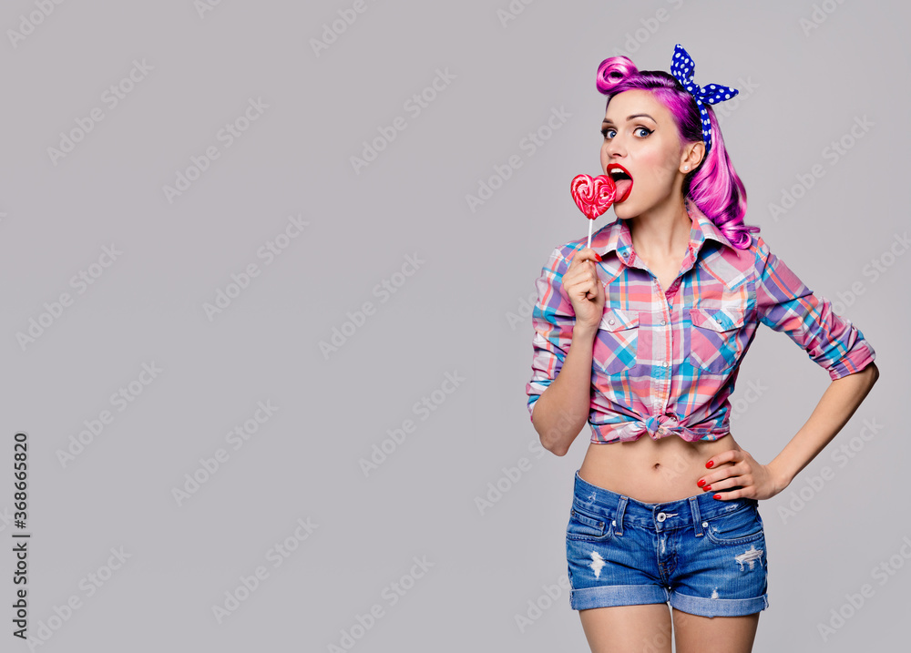 Portrait image of woman licking heart shape lollipop dressed in pinup style. Purple girl in retro fashion and vintage concept. Grey color background. Copy space for some text. Portrait image of woman licking heart shape lollipop dressed in pinup style. Purple girl in retro fashion and vintage concept. Grey color background. Copy space for some text.