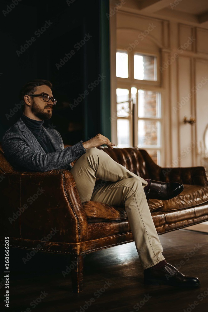 A man in a classic suit sits on a brown leather sofa, in the background a light window A man in a classic suit sits on a brown leather sofa, in the background a light window
