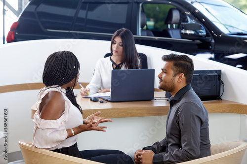 Visiting a car dealership choosing and buying a new car. A young emotional African-American married couple communicate against the background of a car sales agent. Selective focus.
