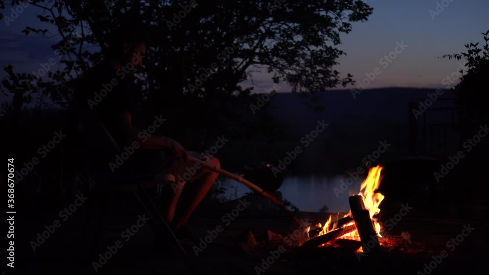 The man straightens the coals in the fire. The fire burns in the night against the background of the river and mountains. The man straightens the coals in the fire. The fire burns in the night against the background of the river and mountains.