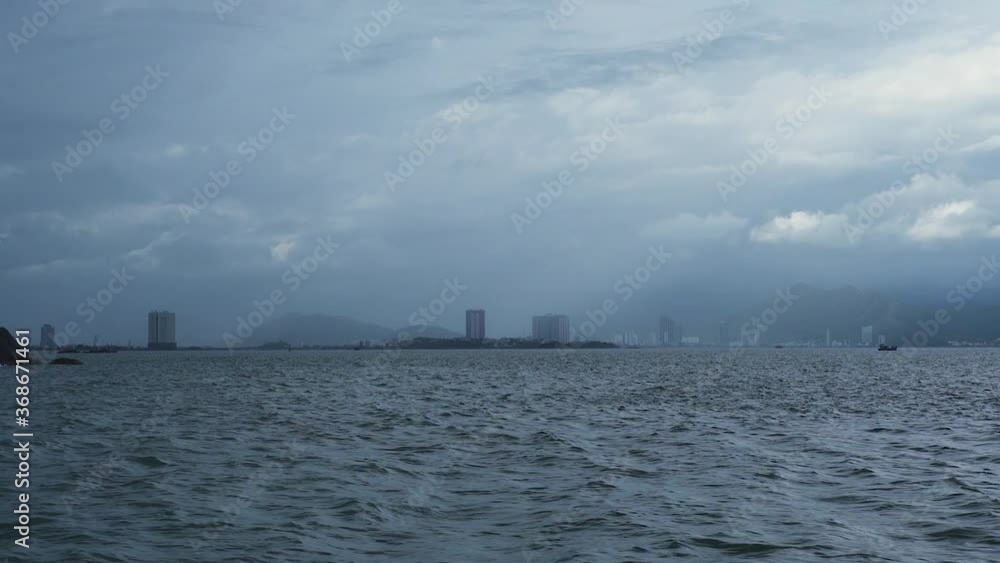 Wavy ocean and dark clouds with view of city buildings and mountains on a moody dramatic day. View from middle of wavy sea, dark clouds over a hazy city and mountains in the distance. Wavy ocean and dark clouds with view of city buildings and mountains on a moody dramatic day. View from middle of wavy sea, dark clouds over a hazy city and mountains in the distance.